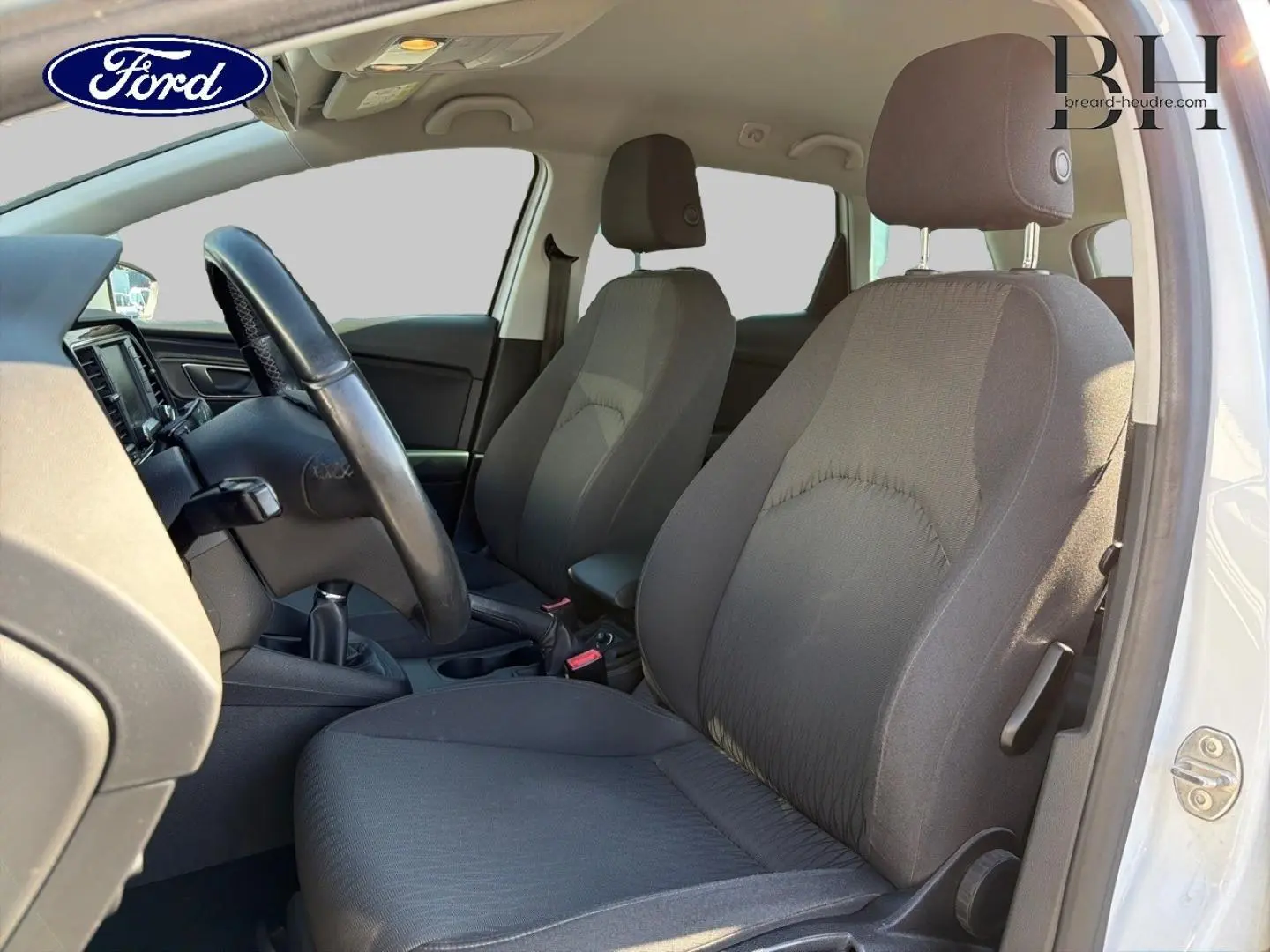 Vue intérieure côté conducteur de la SEAT LEON ST blanche, montrant les sièges tissu noirs et le volant multifonction.