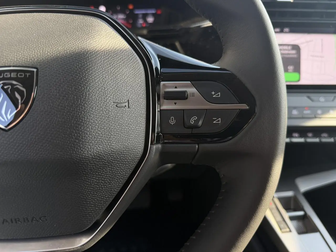 Gros plan sur le volant cuir noir de la Peugeot 308 2025 avec commandes audio et écran tactile en arrière-plan flou.