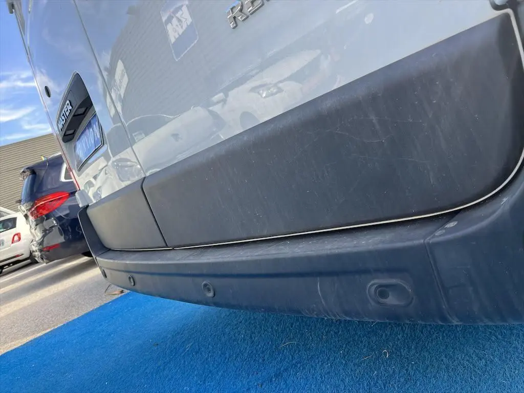 Gros plan sur le pare-chocs arrière noir d'un Renault Master blanc, avec logo et capteurs de recul visibles.