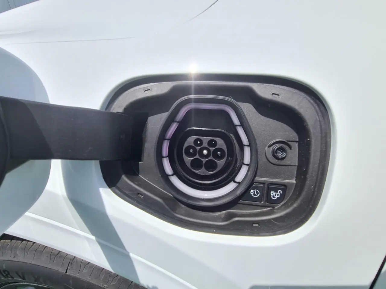 Prise de recharge hybride côté gauche d'un Ford Kuga blanc glacier, avec port de charge ouvert et boutons de commande visibles.