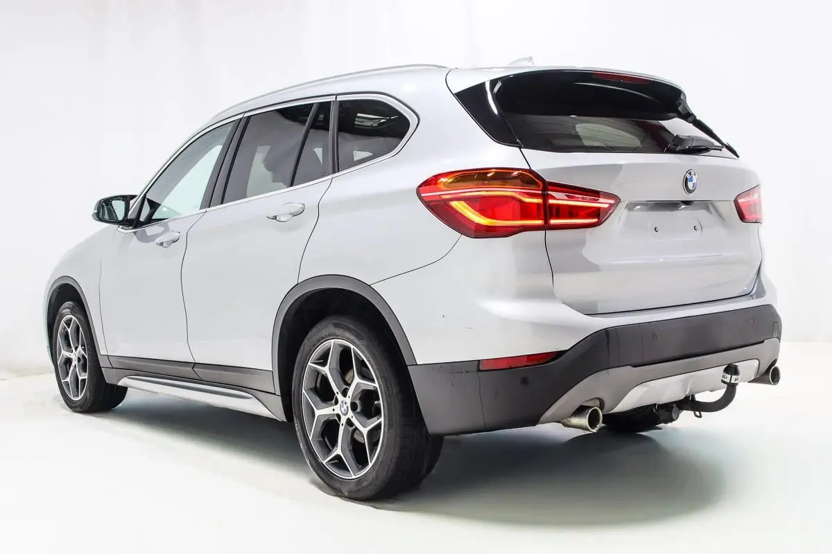 BMW X1 sDrive 18D argent métallisé vue 3/4 arrière droit avec attelage et feux arrière allumés.