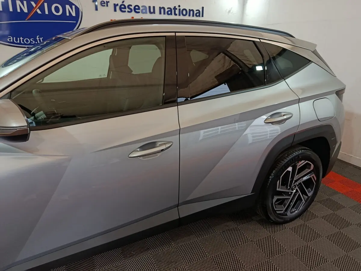 Vue latérale côté gauche d'un Hyundai Tucson 1.6 Hybrid gris clair avec jantes alliage et lignes anguleuses marquées.