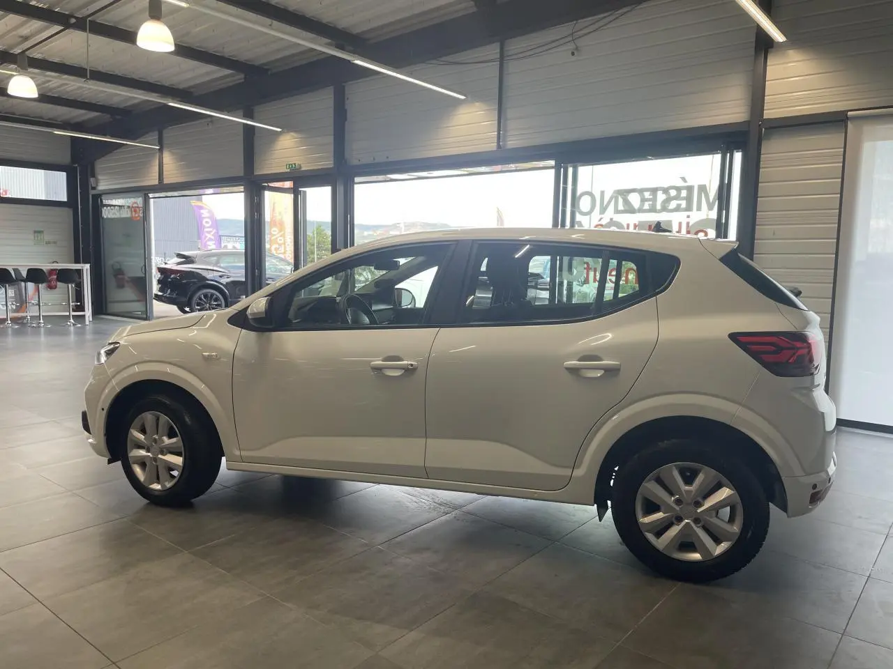 Profil côté gauche d'une Dacia Sandero blanche 2024 en showroom, avec jantes alliage et feux arrière LED distinctifs.