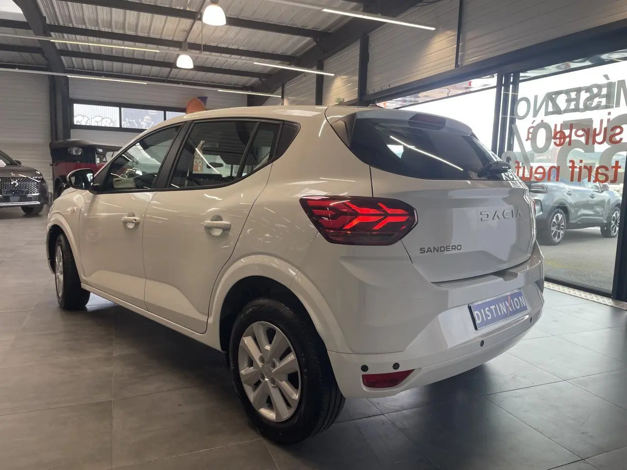 Vue 3/4 arrière droite d'une Dacia Sandero blanche 2024 en showroom, avec feux arrière LED et enjoliveurs 15 pouces.