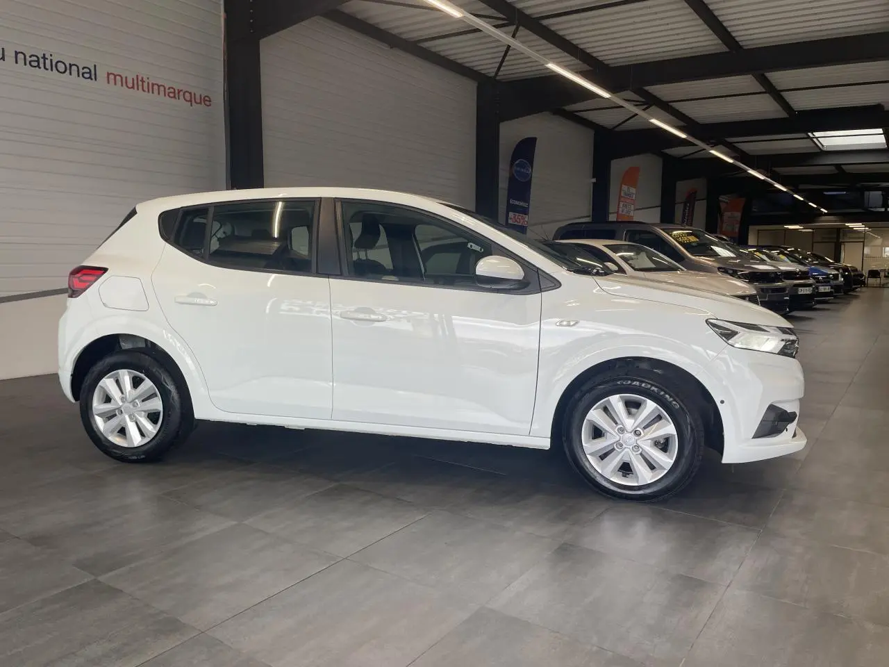 Profil côté gauche d'une Dacia Sandero blanche 2024 en showroom, avec jantes alliage et rétroviseurs couleur carrosserie.