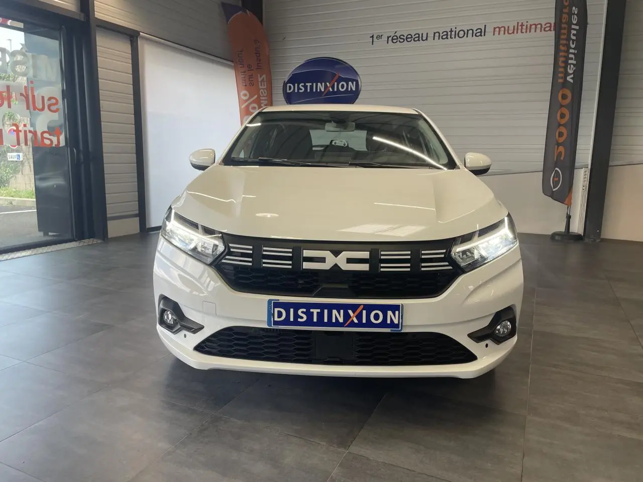 Vue avant d'une Dacia Sandero blanche 2024 en showroom, avec calandre noire et feux LED allumés.