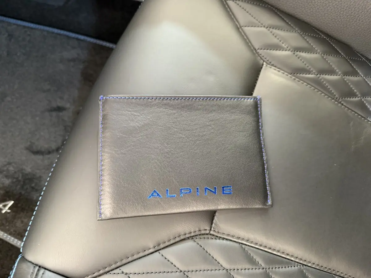 Gros plan sur un porte-documents en cuir noir avec logo Alpine bleu, posé sur un siège en cuir noir matelassé.