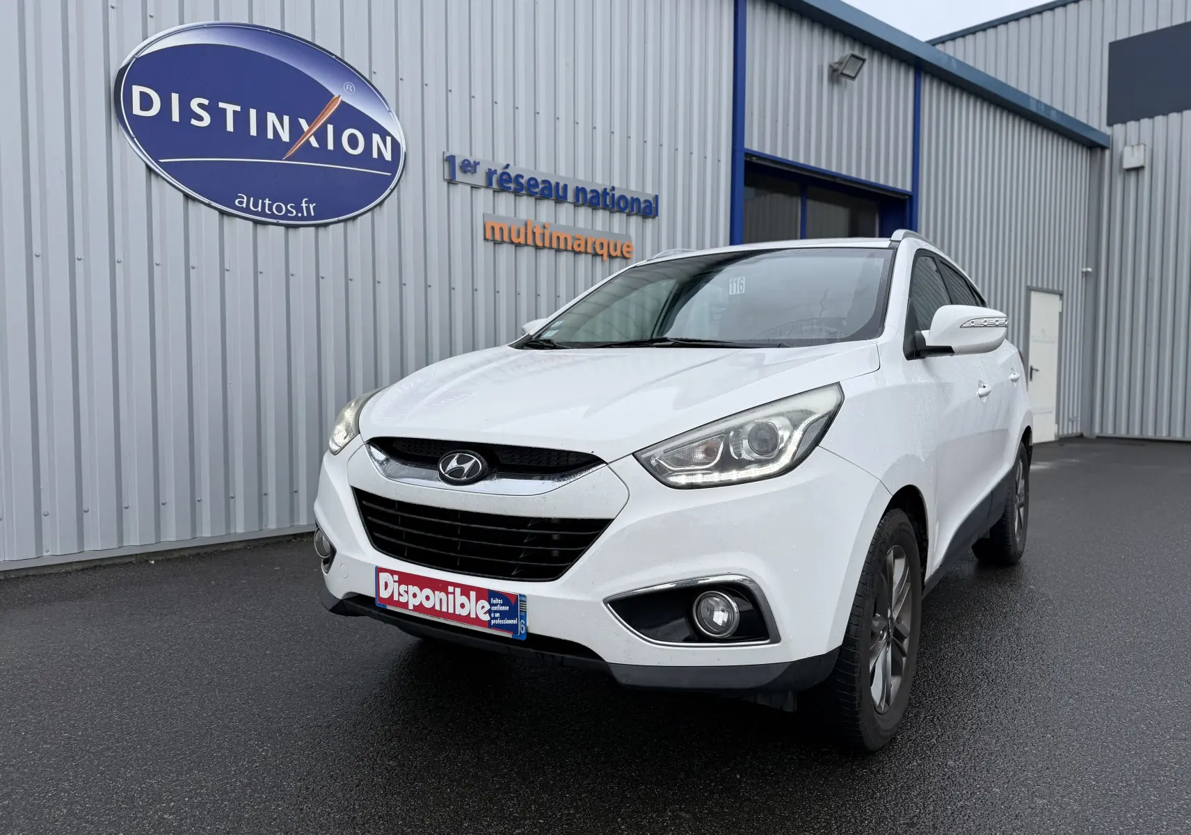 Hyundai ix35 blanc vue 3/4 avant droit, avec phares allumés et plaque "Disponible" devant un bâtiment industriel.