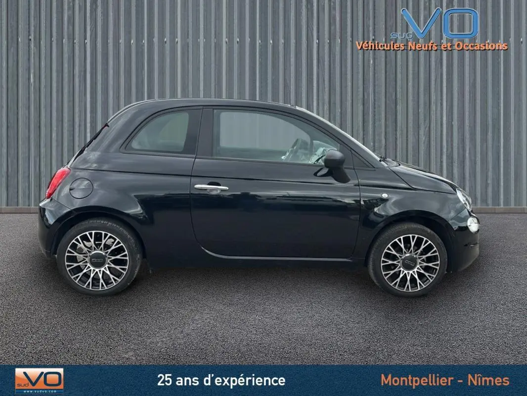 Profil côté gauche de la FIAT 500C noire 2024 avec roues alliage et roue de secours visible sur fond industriel.