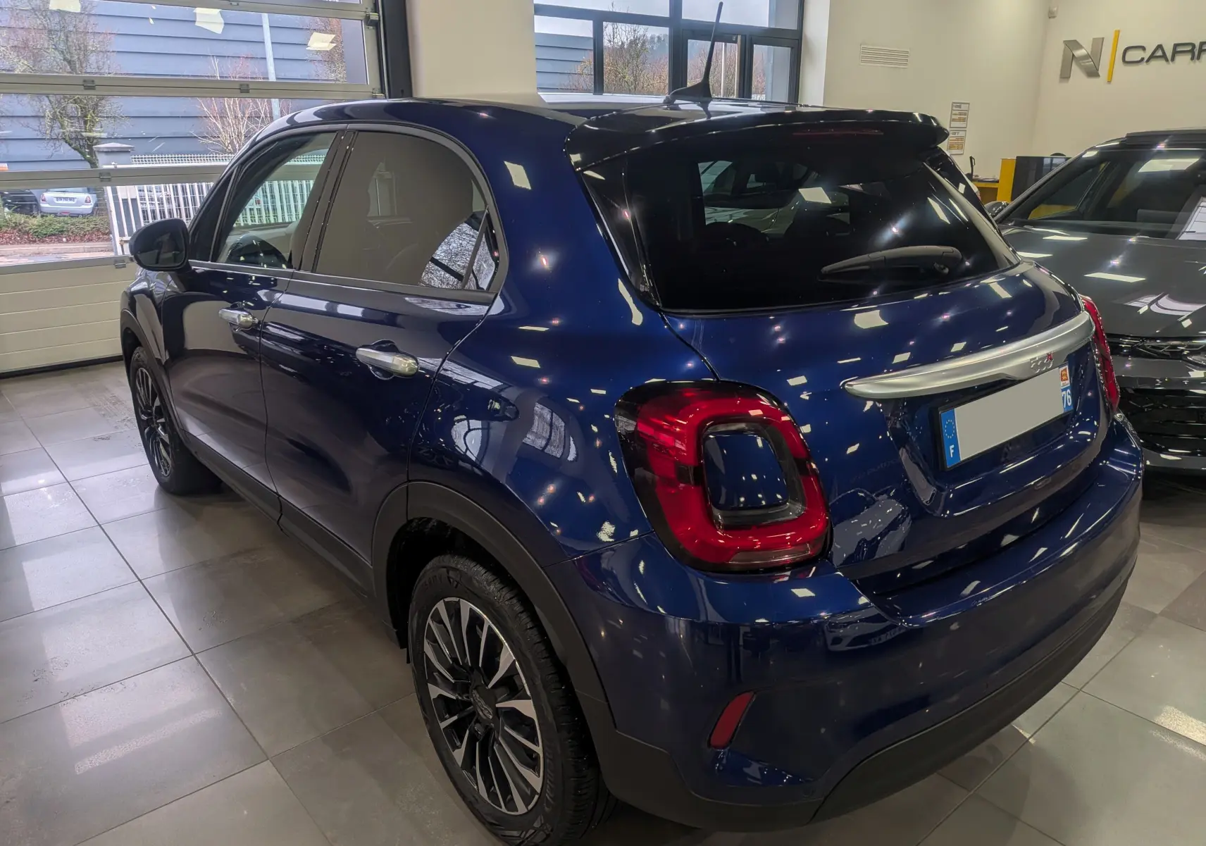 Vue 3/4 arrière droite d'une FIAT 500X 2024 bleu Venezia en intérieur, avec feux arrière distinctifs et jantes noires.
