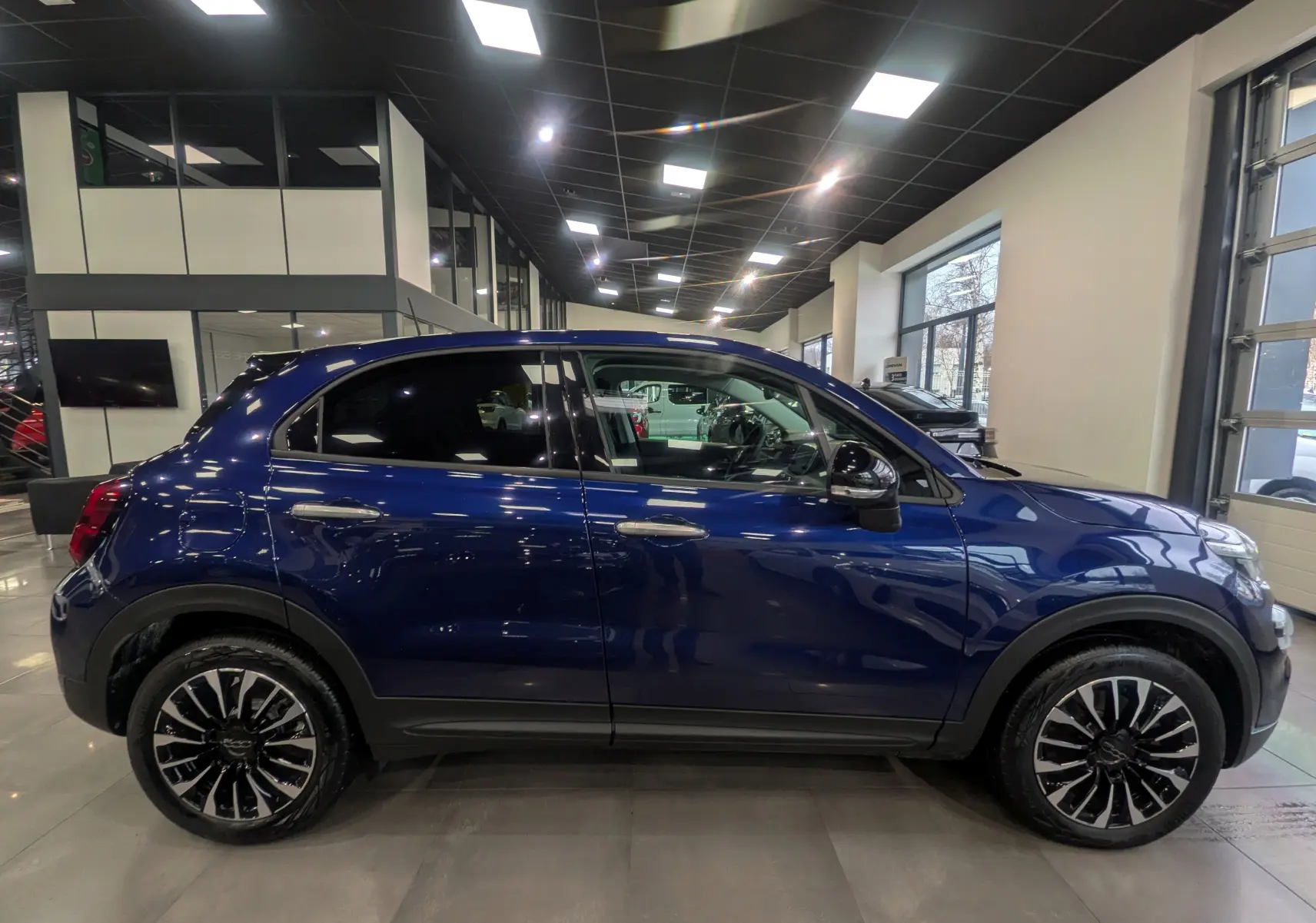 Vue de profil droit d'un FIAT 500X 2024 bleu Venezia avec jantes alliage noires et toit noir en concession.