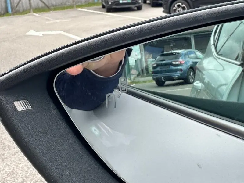 Reflet dans le rétroviseur côté gauche d'un SEAT Arona blanc Nevada avec toit noir, montrant une partie du parking.