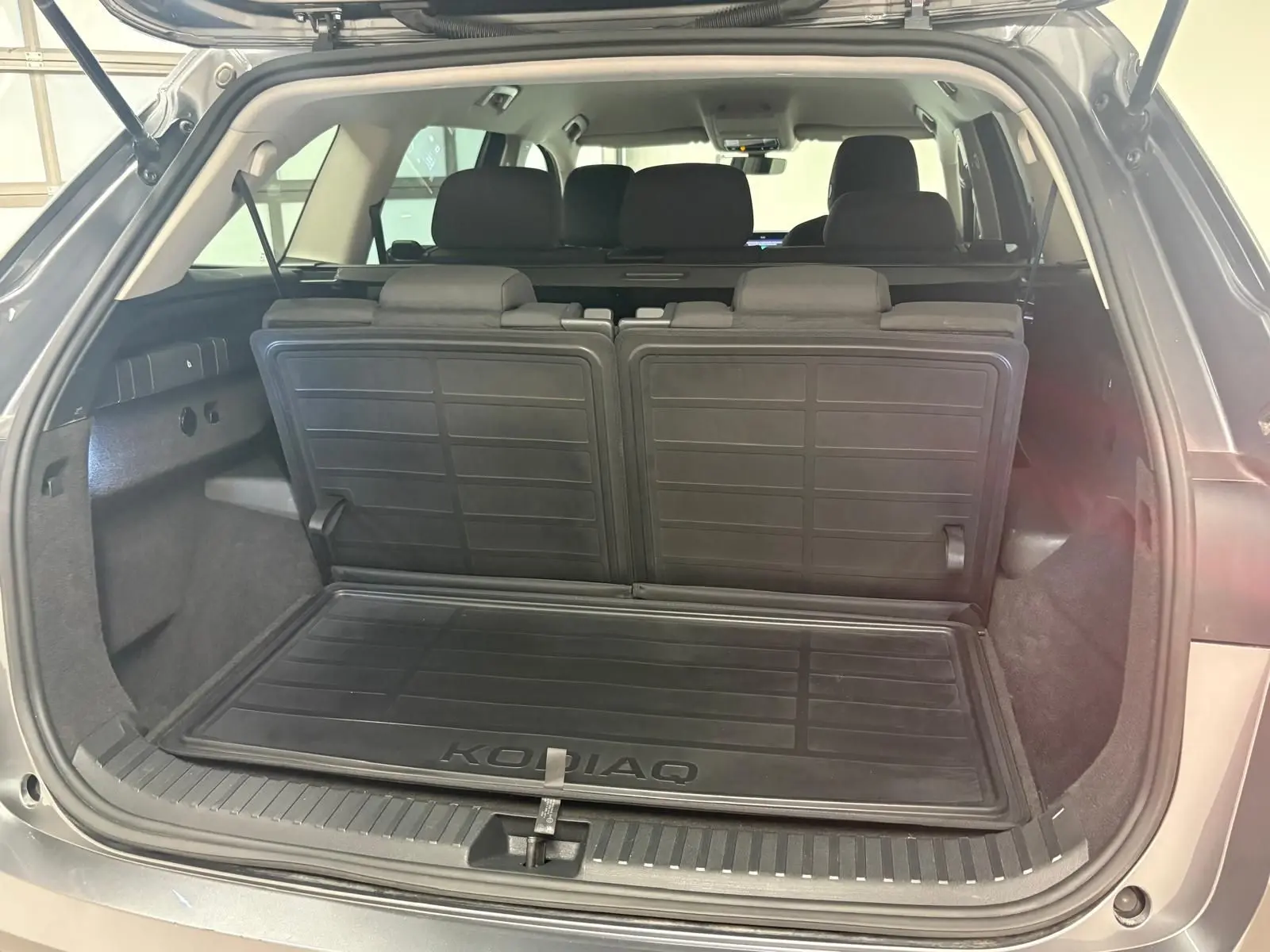 Vue arrière du coffre ouvert du Skoda Kodiaq gris graphite 2025, montrant la banquette rabattue et le tapis de coffre marqué KODIAQ.