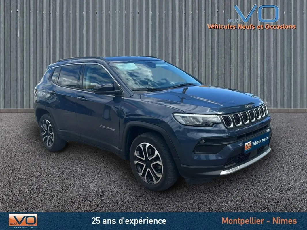 Jeep Compass bleu 3/4 avant droit avec barres de toit et jantes 18 pouces, stationné devant un mur métallique.