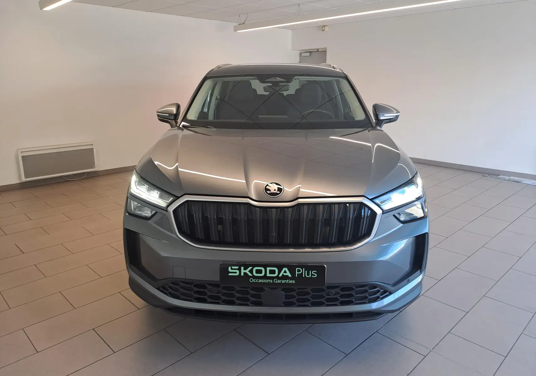 Vue avant d'un Skoda Kodiaq gris graphite 2025 avec calandre noire et phares allumés en intérieur showroom.