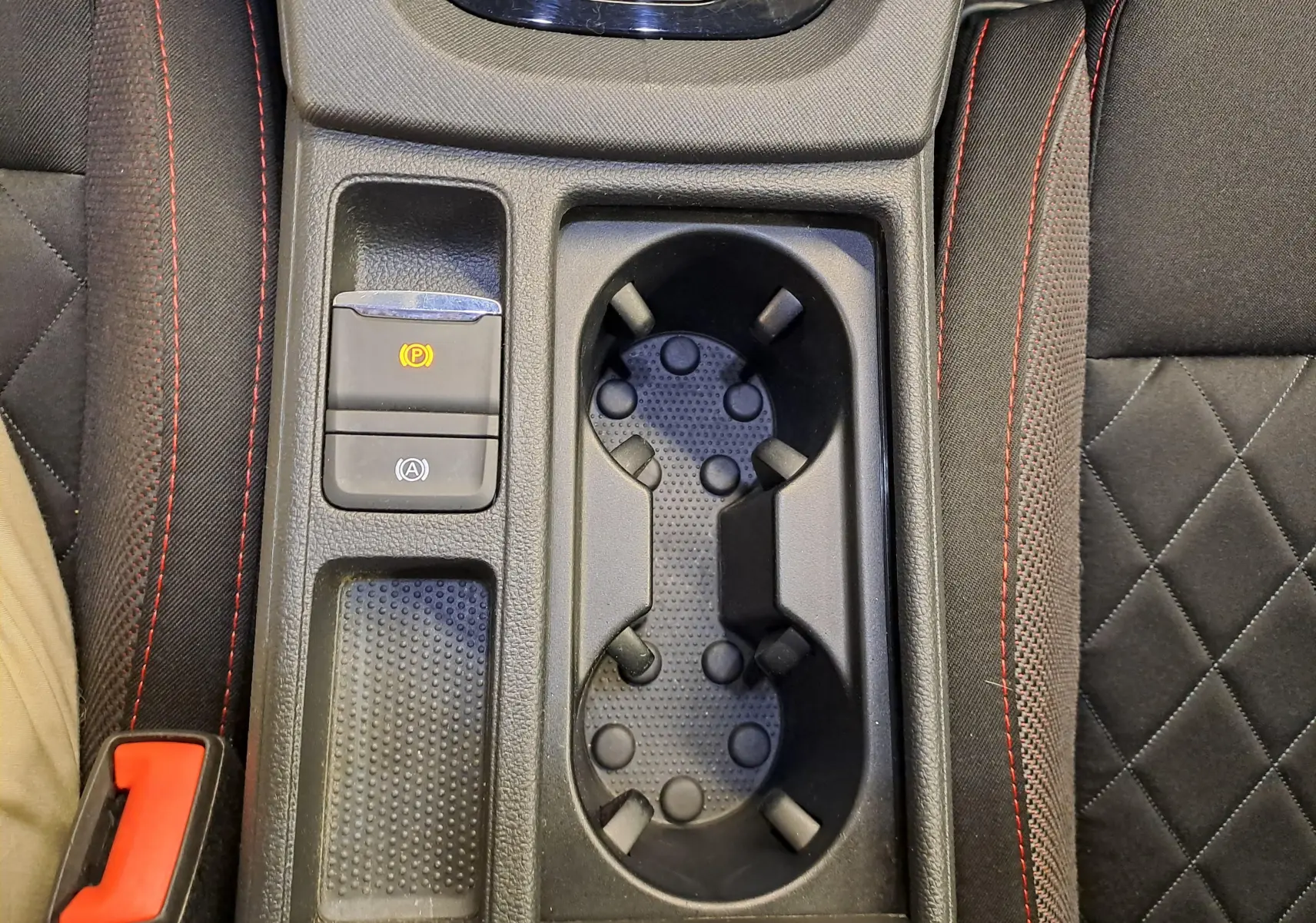 Gros plan sur la console centrale avec porte-gobelets et bouton frein à main électrique, tissu noir avec surpiqûres rouges.