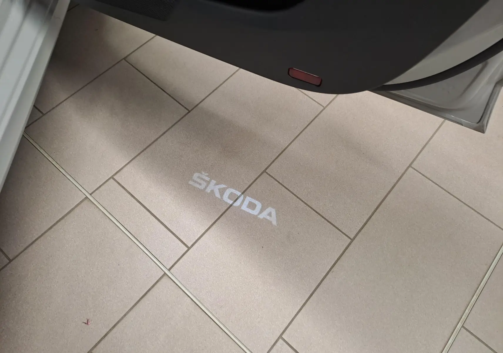 Projection lumineuse du logo ŠKODA au sol vue depuis la porte ouverte côté gauche d’un véhicule gris perle.