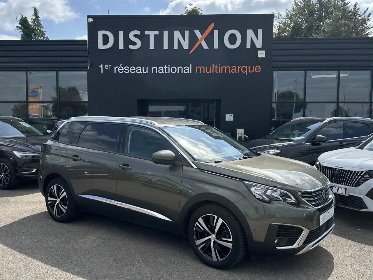 Peugeot 5008 gris amazonite en 3/4 avant droit, avec jantes alliage et rétroviseurs chromés, stationné devant un showroom.
