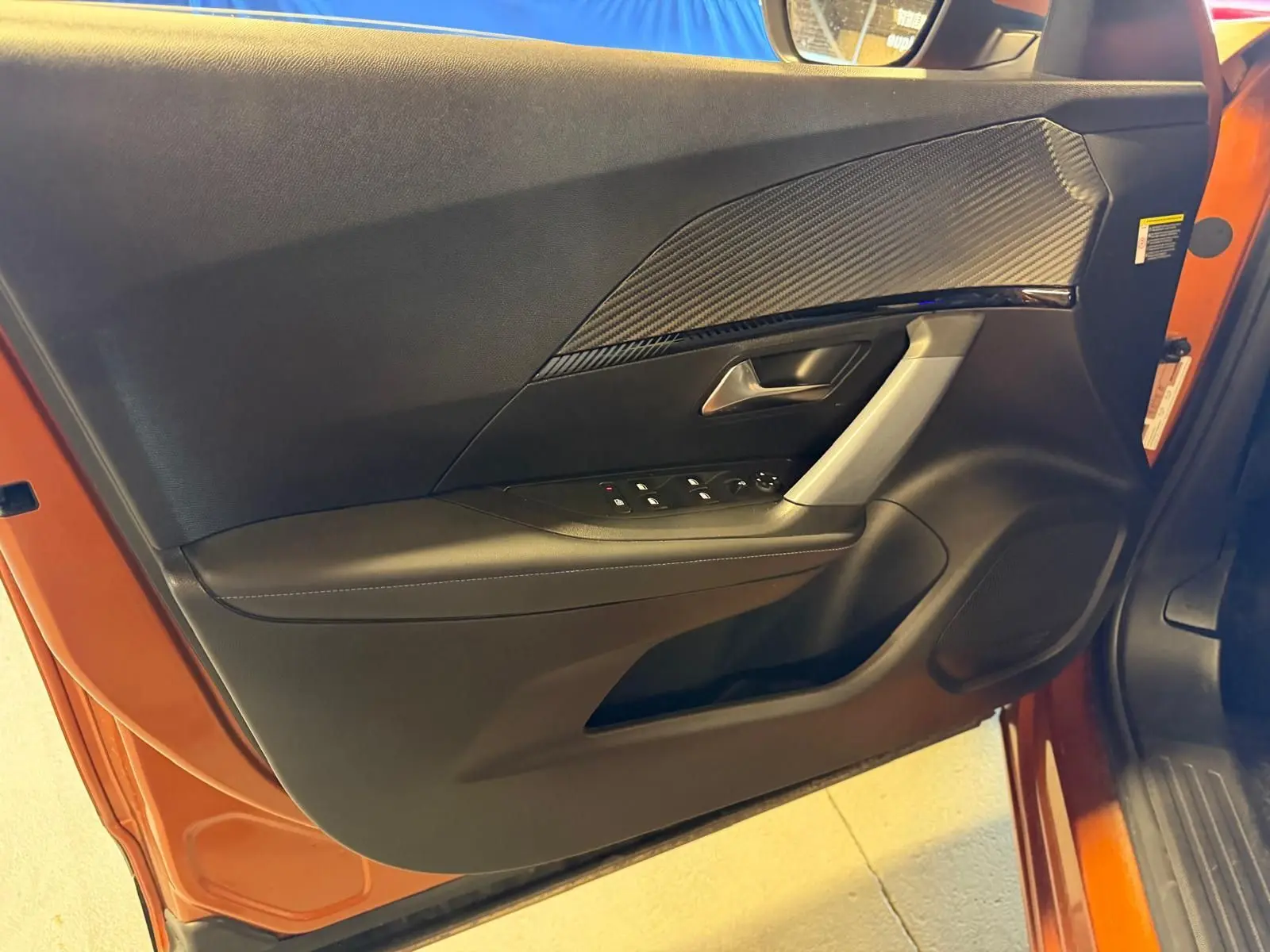 Vue intérieure de la porte avant gauche orange du Peugeot 2008 1.5 BlueHDi avec insert carbone et commandes électriques.