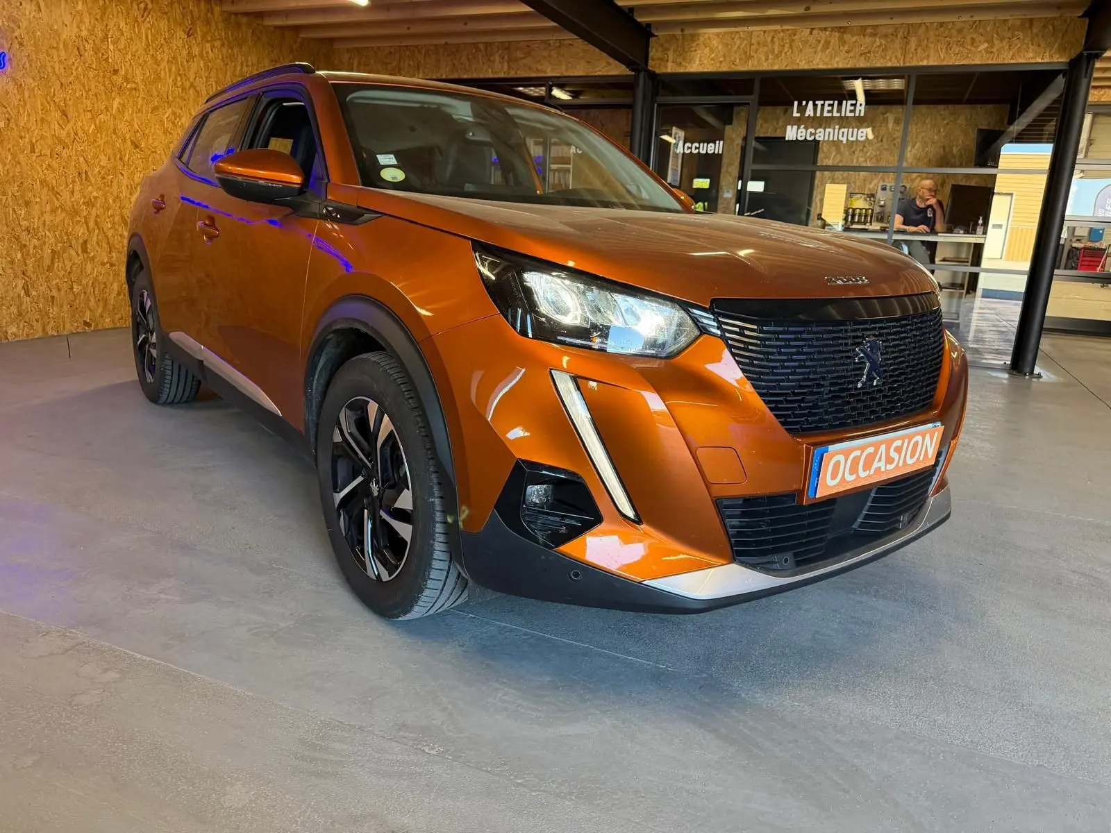 Peugeot 2008 orange vue 3/4 avant droit, avec calandre noire et jantes alliage bicolores, en intérieur showroom.