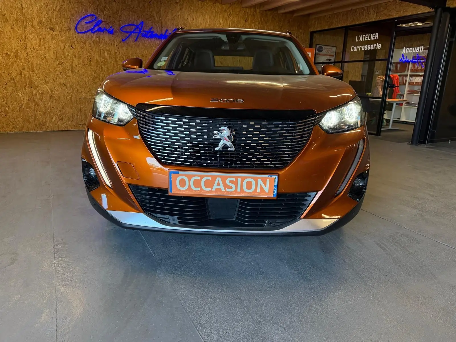 Vue de face d'un Peugeot 2008 orange métallisé avec calandre noire et phares LED allumés en showroom.