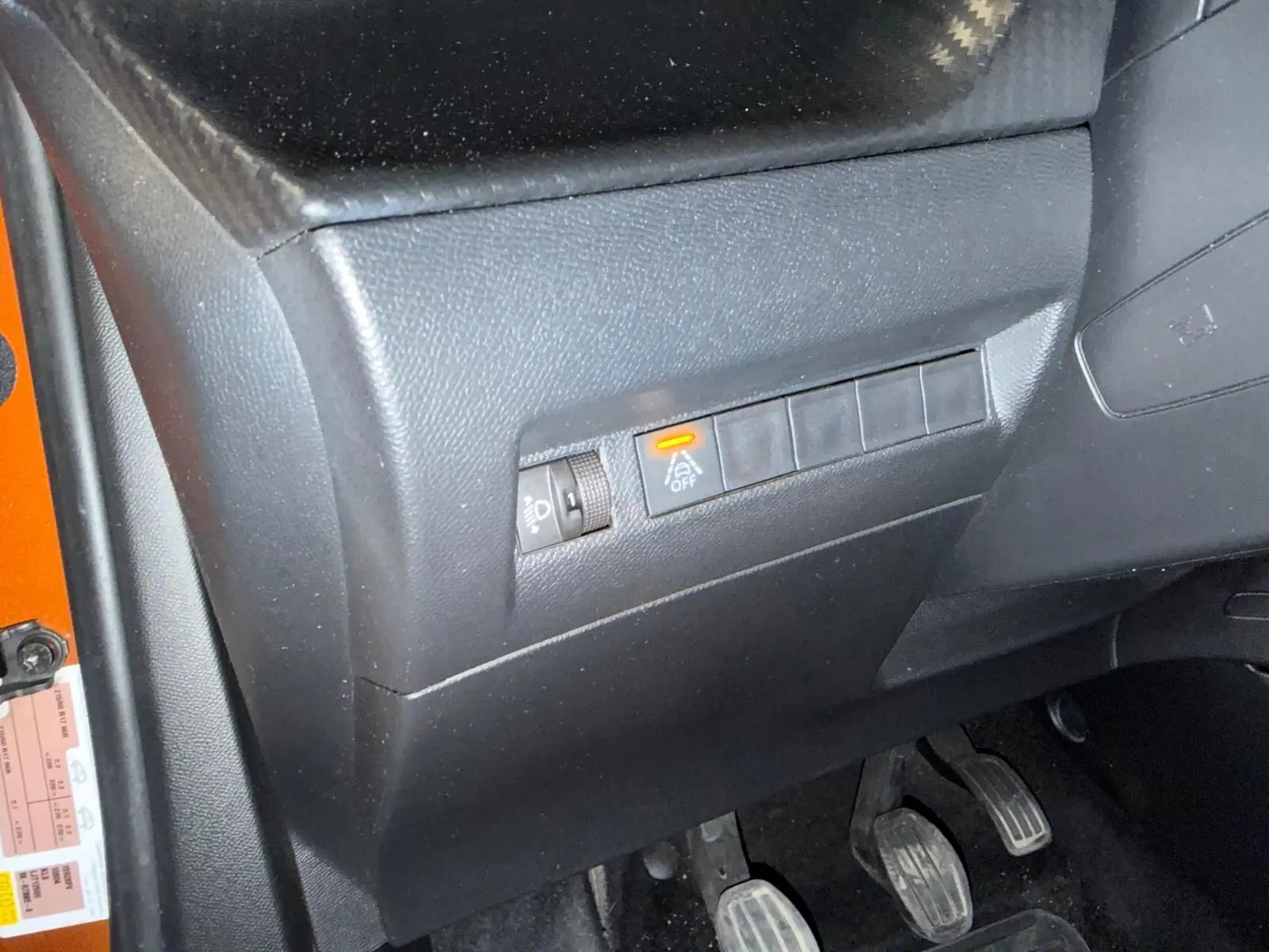Détail du tableau de bord côté conducteur d’un Peugeot 2008 orange, avec commande d’assistance désactivée et pédales visibles.