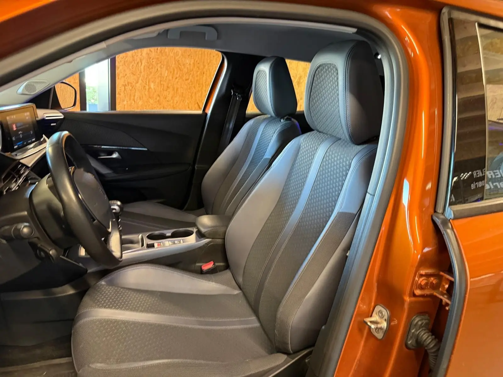 Vue intérieure côté conducteur de la Peugeot 2008 orange, montrant sièges tissu gris et volant compact cuir.