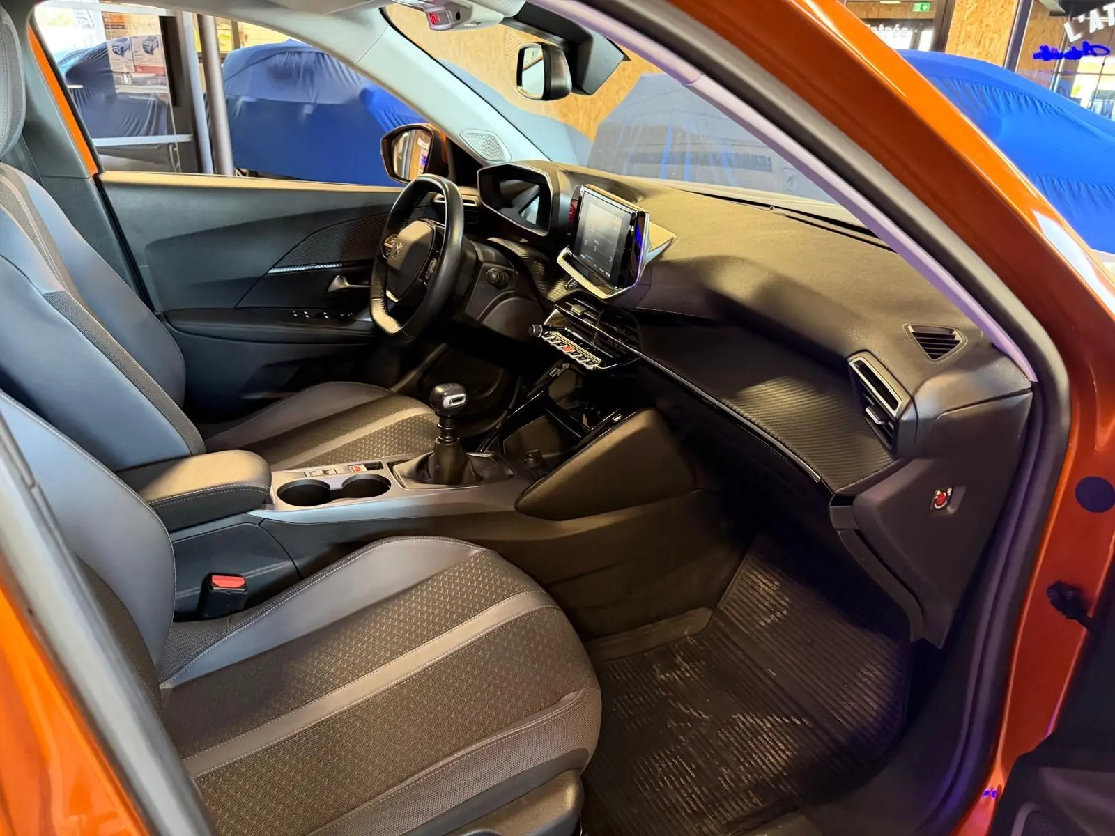 Vue intérieure côté conducteur du Peugeot 2008 orange, montrant la planche de bord et la sellerie tissu grise avec surpiqûres.
