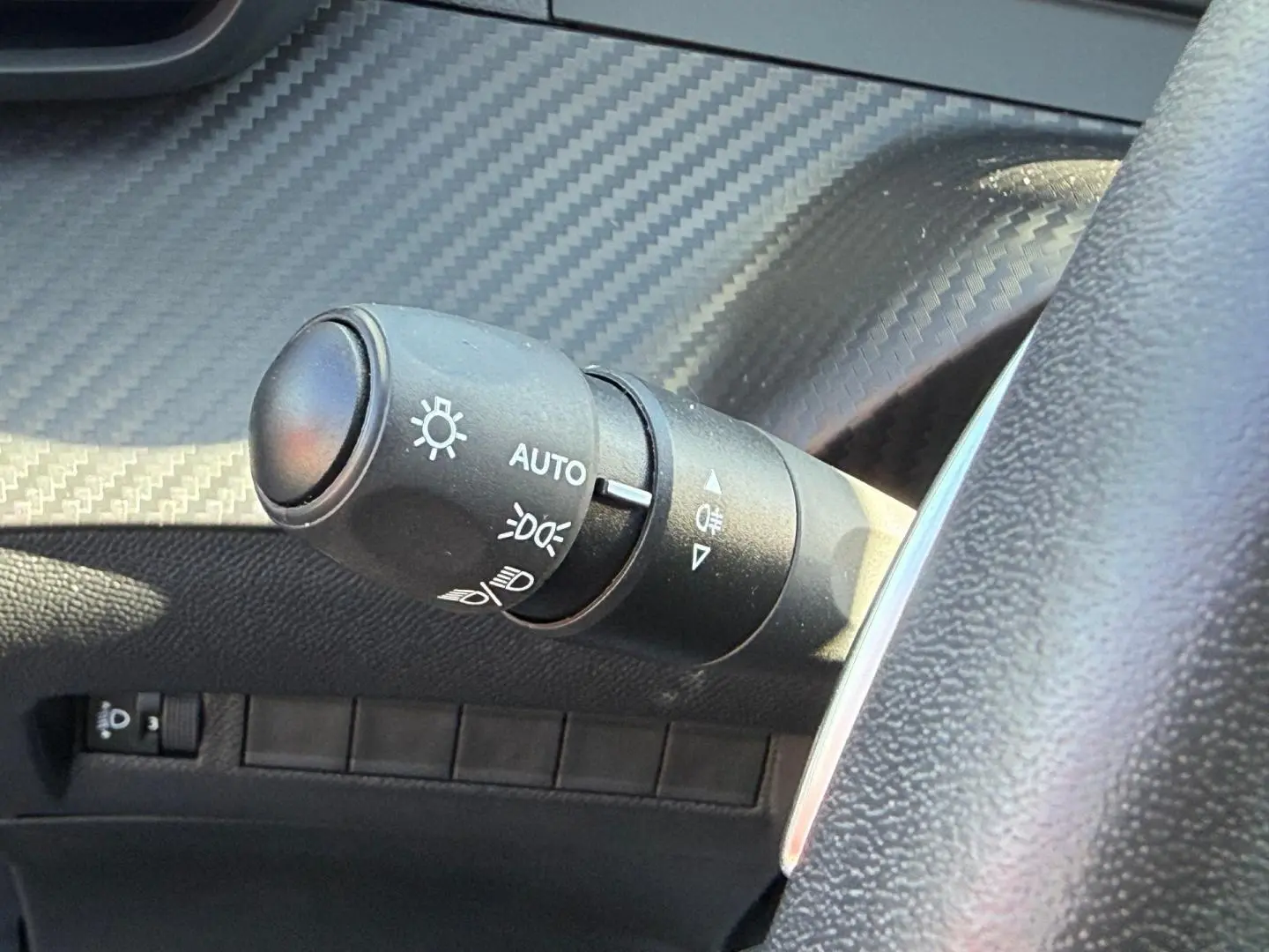 Gros plan sur la commande des phares avec finition effet carbone et volant partiel gris dans une Peugeot 208 gris Artense.