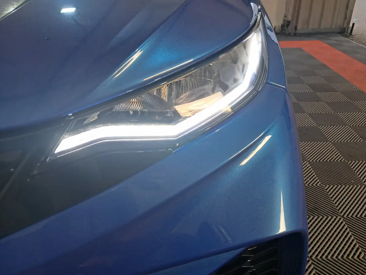 Gros plan sur le phare avant droit d'une Aixam Ambition 2025 bleu métallisé avec éclairage LED allumé.