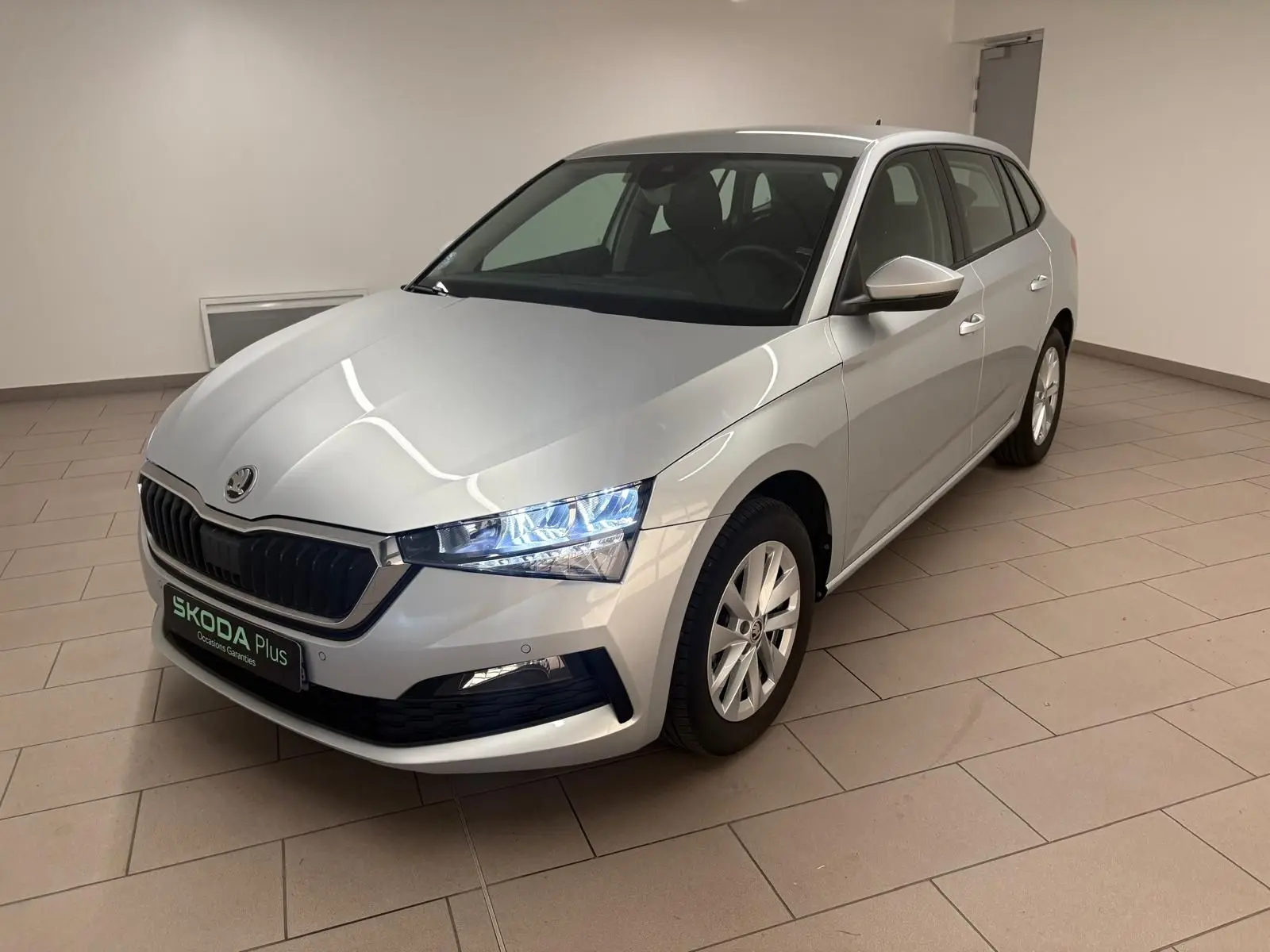 Vue 3/4 avant droite d'une Skoda Scala gris argent 2024 avec phares allumés dans un intérieur neutre.