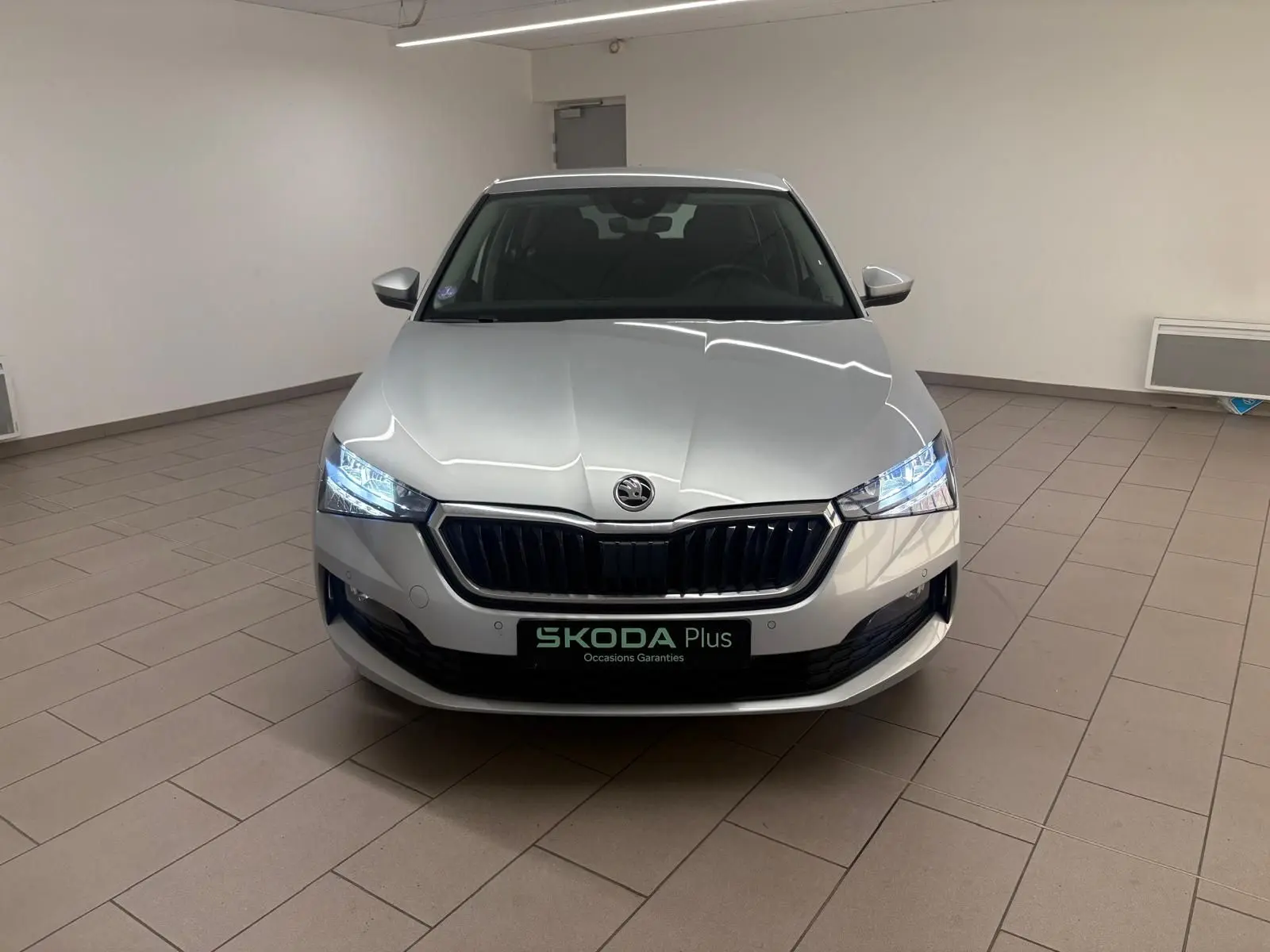 Vue frontale d'une Skoda Scala 1.0 TSI 110 DSG7 Business gris argent avec phares allumés en intérieur showroom.