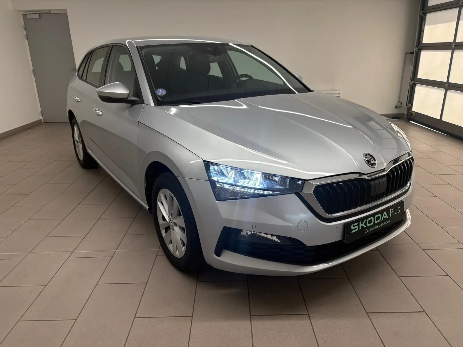 Vue 3/4 avant droite d'une Skoda Scala gris argent 2024 avec phares allumés dans un showroom intérieur.