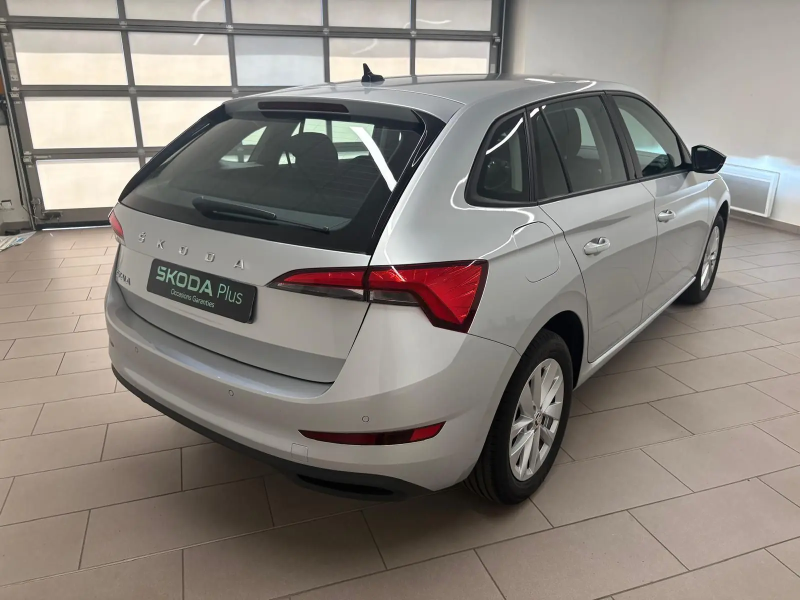Vue 3/4 arrière droite d'une Skoda Scala gris argent 2024 en intérieur, avec feux arrière rouges et jantes argentées.