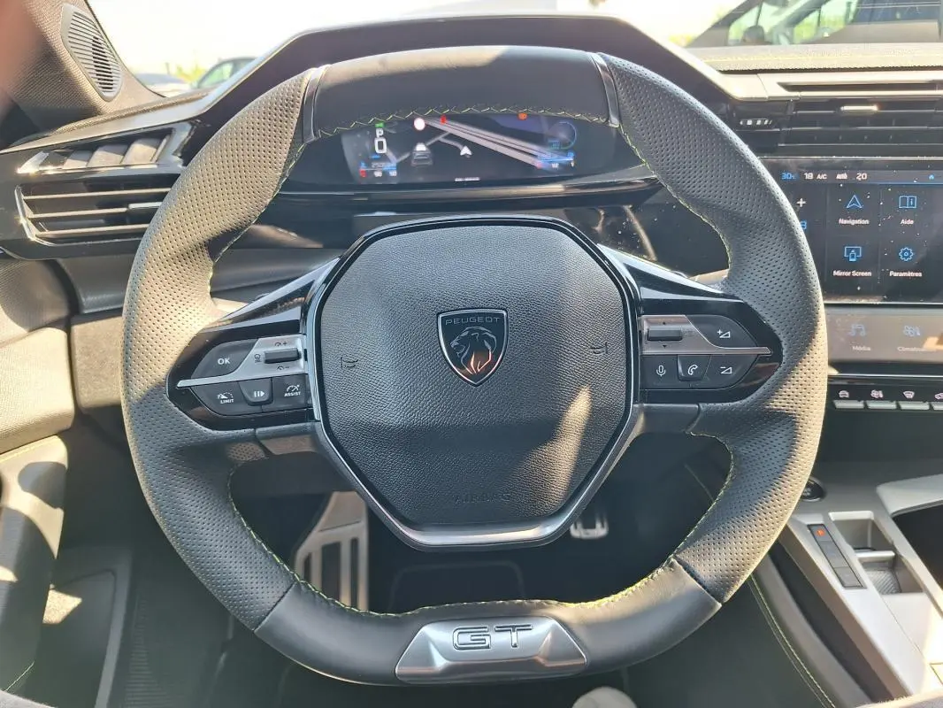 Vue rapprochée du volant cuir noir avec logo Peugeot et badge GT, tableau de bord numérique et écran tactile en arrière-plan.