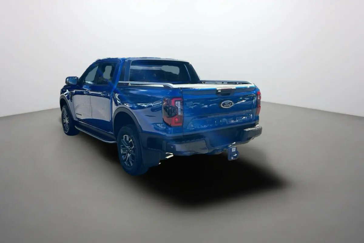 Ford Ranger 2.0 Wildtrak Plus bleu lightning vu 3/4 arrière droit avec attelage et barres de toit visibles.