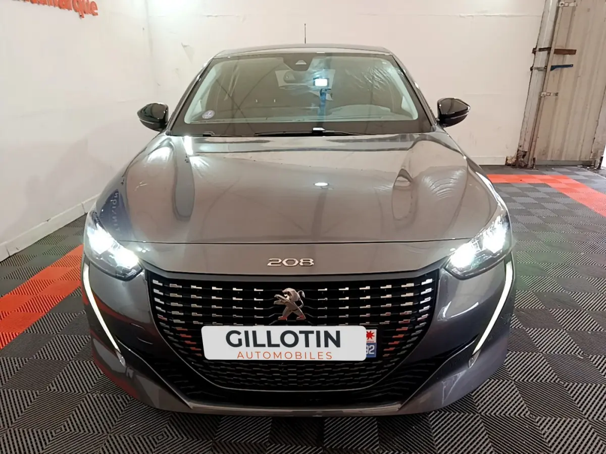 Vue frontale d'une Peugeot 208 gris foncé avec phares LED allumés dans un garage.