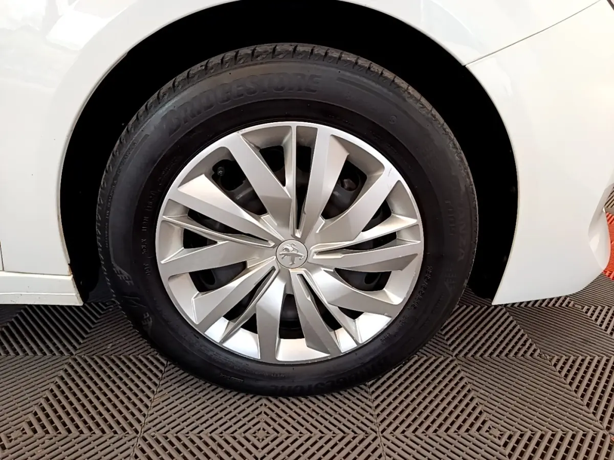 Gros plan sur la roue avant gauche d'une Peugeot 208 blanche, mettant en valeur la jante en alliage au design moderne.