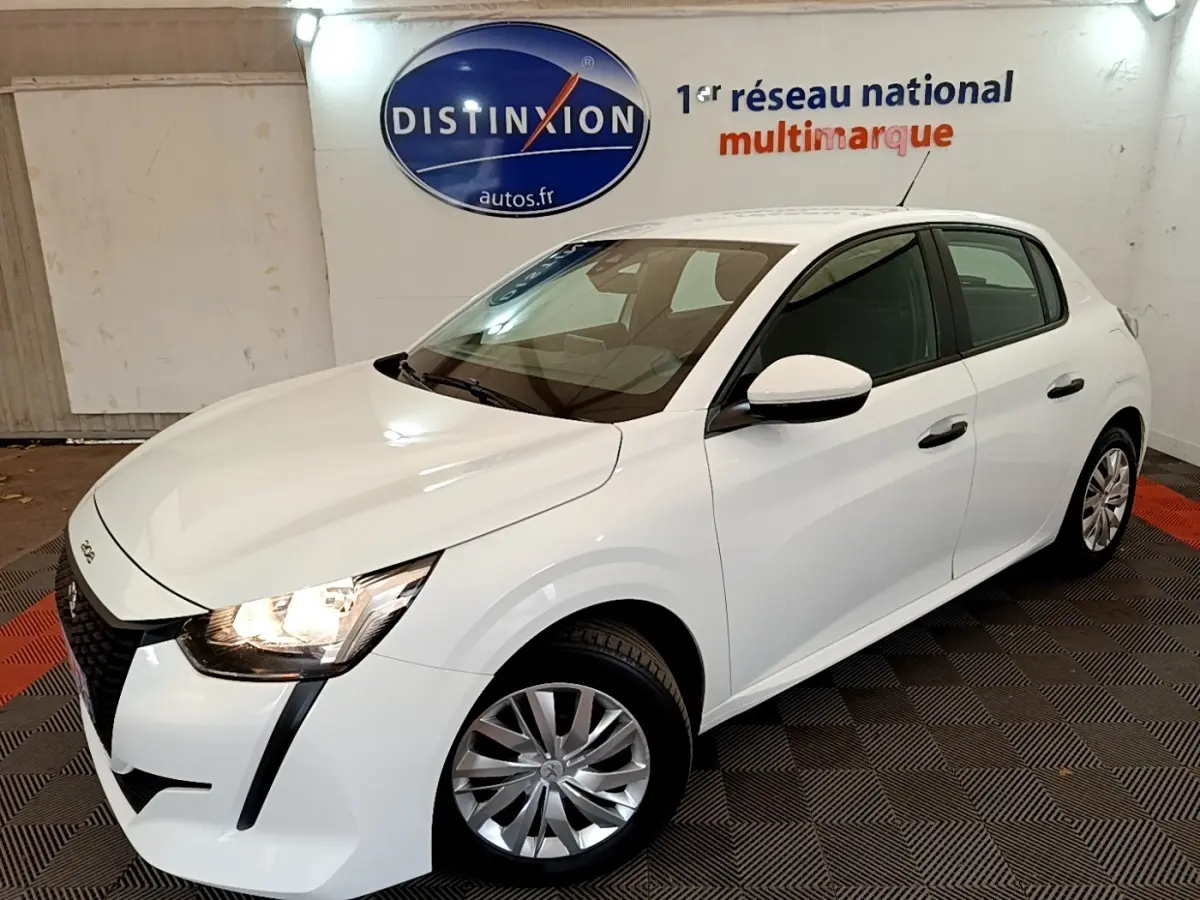 Peugeot 208 blanc vue 3/4 avant droit en intérieur, avec phares allumés et jantes alliage visibles.