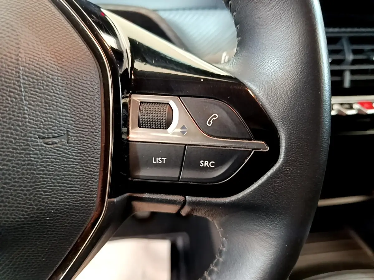 Gros plan sur les commandes gauche du volant en cuir noir d'une Peugeot 208 blanche, avec boutons de téléphone et liste.