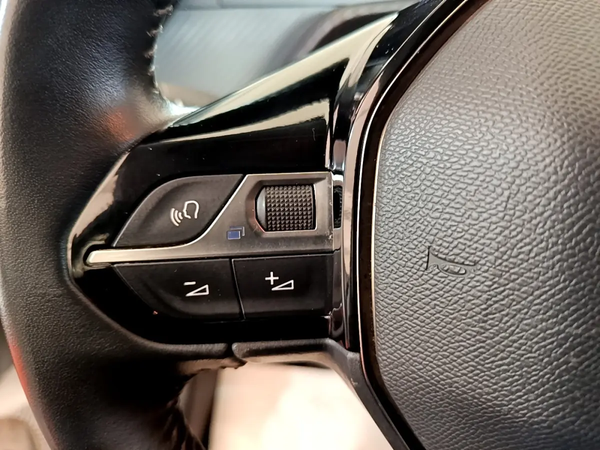 Gros plan sur les commandes gauche du volant noir de la Peugeot 208 blanche, avec boutons de volume et appel vocal.