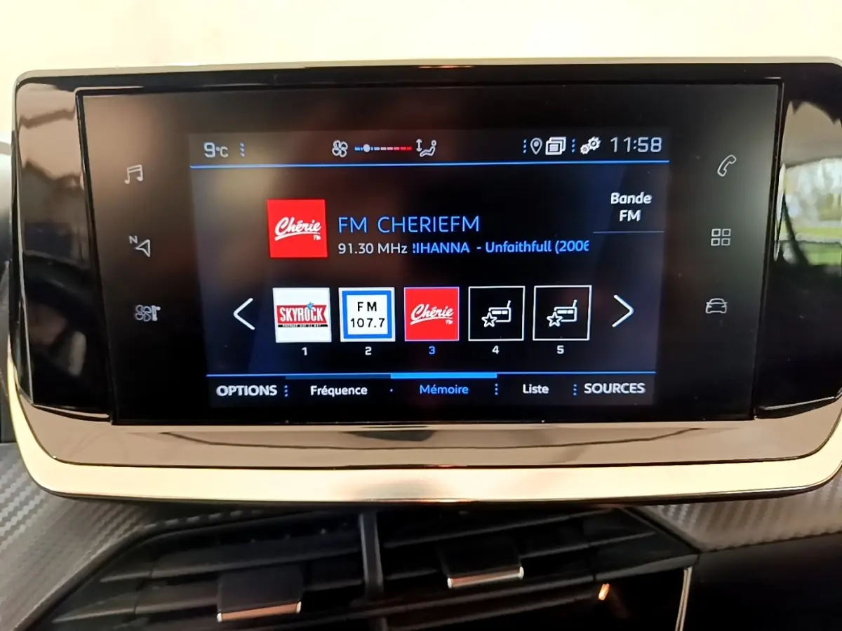 Écran tactile central affichant la radio FM dans l'habitacle d'une Peugeot 208 blanche, vue de face.