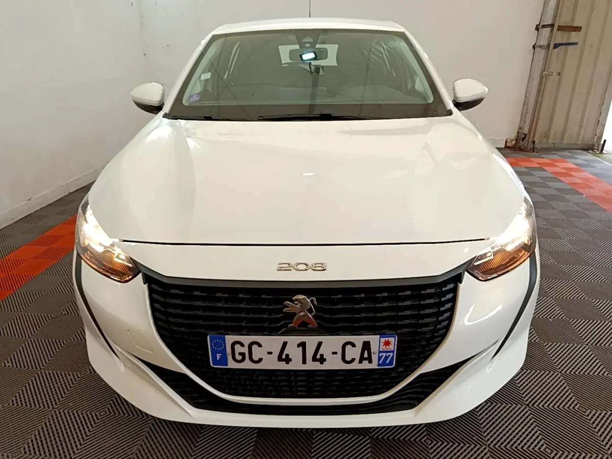 Vue avant d'une Peugeot 208 blanche avec calandre noire et phares allumés dans un garage.