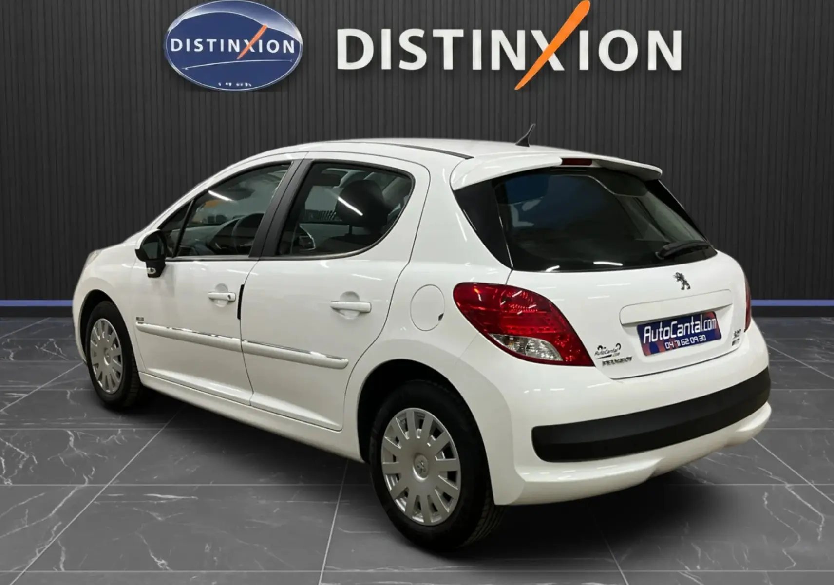 Vue 3/4 arrière droite d'une Peugeot 207 blanche avec baguettes chrome et béquet, stationnée en intérieur showroom.