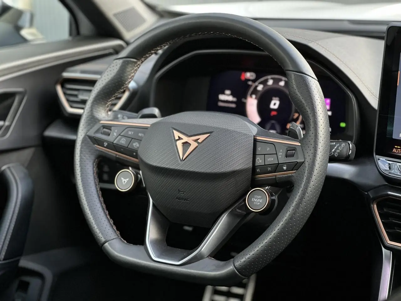 Vue rapprochée du volant cuir perforé noir avec logo CUPRA cuivré et commandes intégrées, tableau de bord numérique en arrière-plan.