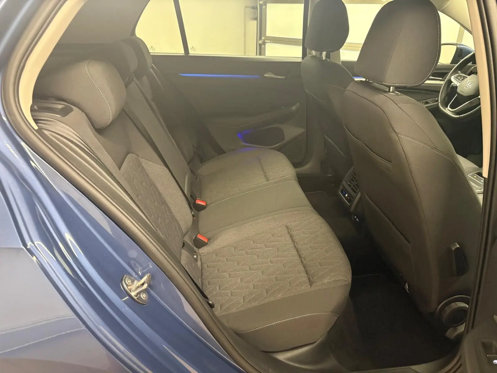 Vue intérieure arrière côté gauche de la Volkswagen Golf 1.5 TSI 2025 en Bleu Anémone, sièges tissu gris et éclairage d'ambiance bleu.