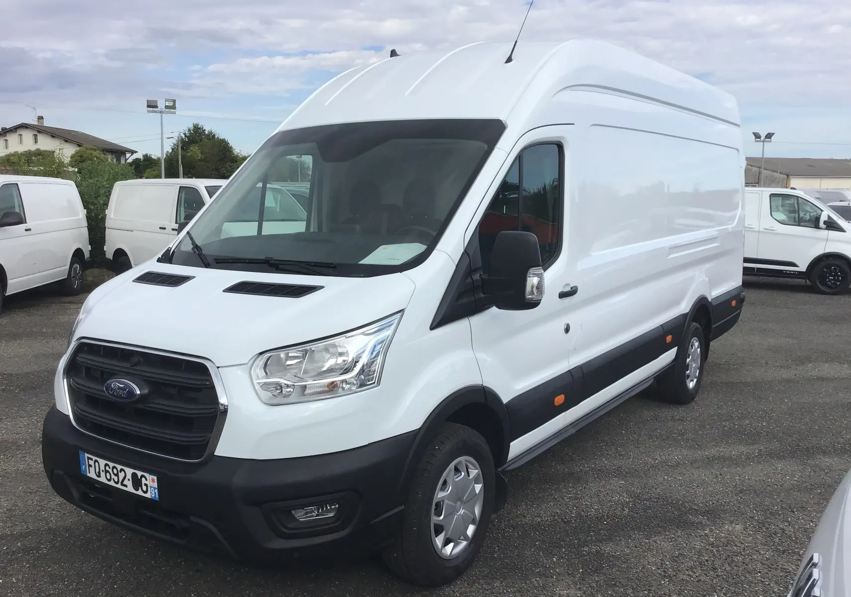 Fourgon Ford Transit blanc en vue 3/4 avant droit, avec calandre noire et rétroviseurs noirs sur parking.