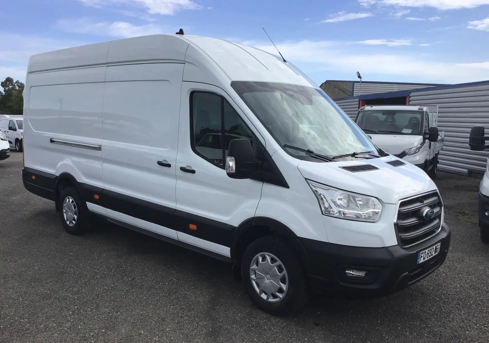 Ford Transit Fourgon blanc en 3/4 avant droit, avec toit haut et pare-chocs noirs visibles sur un parking extérieur.