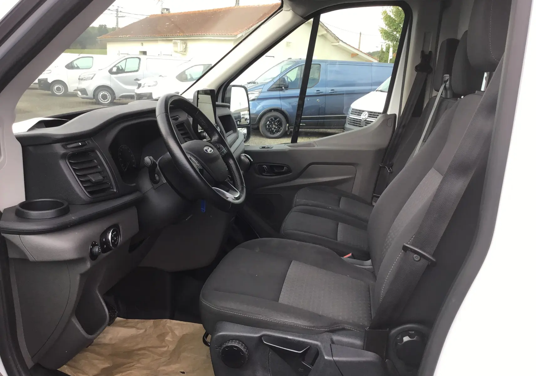 Vue intérieure côté conducteur du Ford Transit Fourgon blanc 2020, montrant le tableau de bord et les sièges noirs.