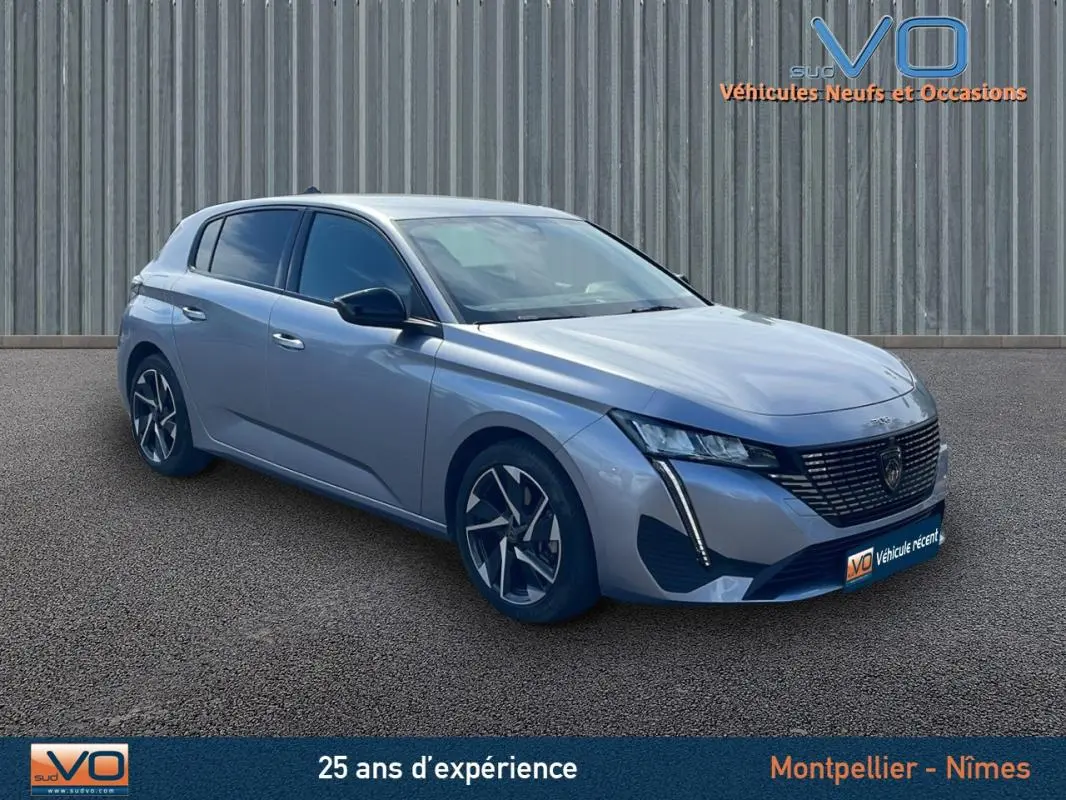 Peugeot 308 gris 3/4 avant droit, avec calandre distinctive et jantes modernes, stationnée devant un mur métallique.